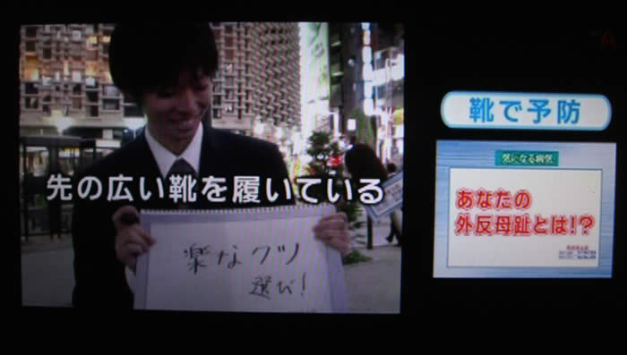 (番組キャプチャ)画面右側に 靴で予防 あなたの外反母趾とは!?と表示されている。画面左には、夜の駅前でスーツを着た若い男性が「楽なクツ選び!」と書いたバードを持っており、その上からテロップが 先の広い靴を履いている と表示