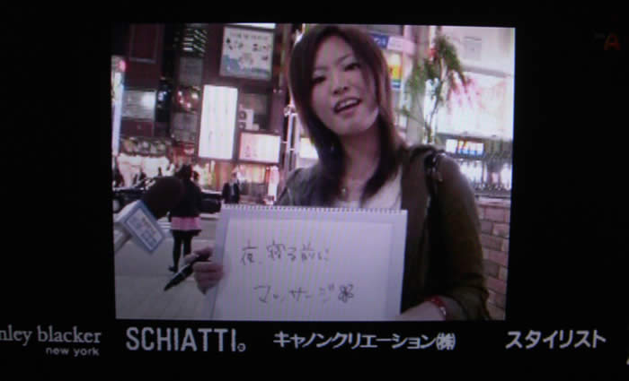 (番組キャプチャ)画面中央に、夜の駅前で普段着を着た若い女性が「夜寝る前にマッサージ🌼」と書いたバードを持っている。
