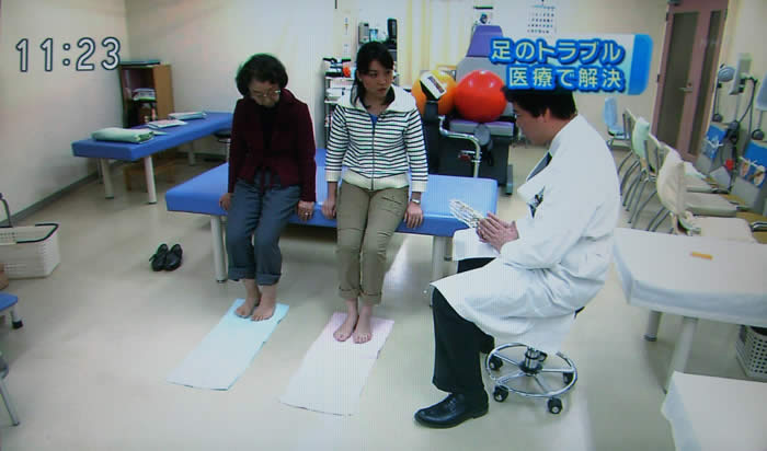 (番組キャプチャ)診察台に腰掛ける患者さんとキャスター、その横で吉野院長が足の骨の模型を手に説明をしている